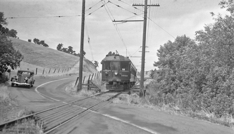 A Sacramento Northern electric interurban train rounds a bend on the Shepherd Canyon corridor, mid-twentieth century. A vintage car shares the parallel road.